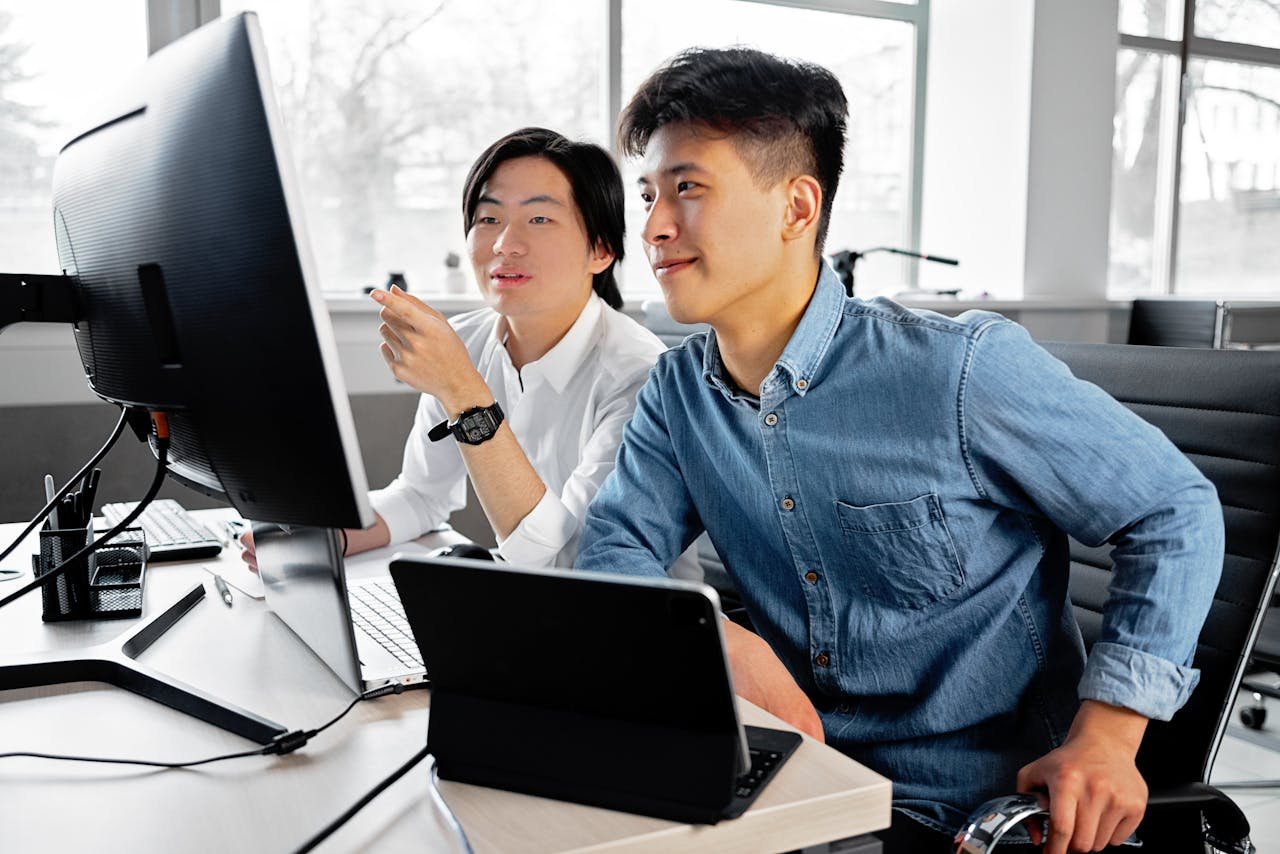 Two men collaborating on a project at a desktop computer in a bright office setting.